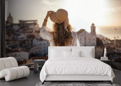 Blonde woman standing on the balcony and looking at coast view of the southern european city with sea during the sunset, wearing hat, cork bag, safari shorts and white shirt Wall mural