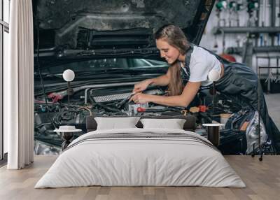 A brunette in a black jumpsuit and a white t-shirt near the open hood of black car. Young female in the garage is smiling at the camera and lowered gaze. car repair concept Wall mural