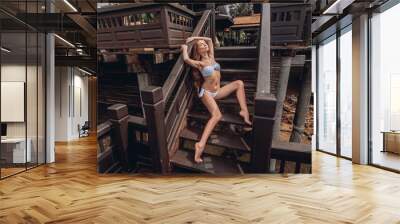 A young beautiful female with long curly brown hair in a stylish white bikini poses sitting on the stairs of a wooden luxury villa. Summer tropical photosession. Wall mural