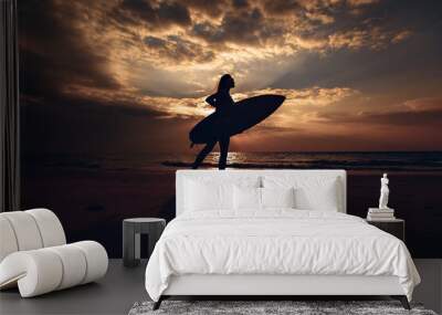 Silhouette of the surfer slim girl with surfboard in her hand on the sandy beach at sunset. Surfer and ocean. Side view. Wall mural