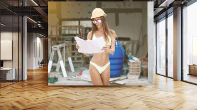 Very hot and bright girl in a bikini, helmet and glasses is watching the construction of the building, holding the project in her hands, portrait Wall mural