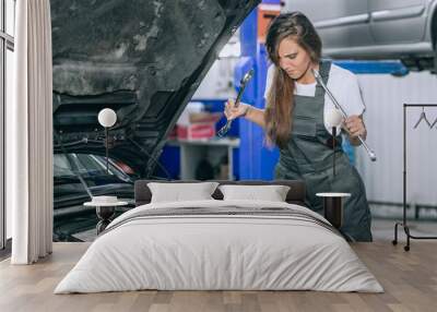 Young brunette near the open hood of a black car in the garage. The girl is dressed in black jumpsuit and white t-shirt, she holds up the wrenches and lowers her gaze. Wall mural