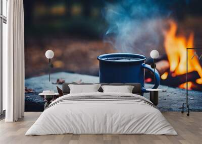 blue enamel cup of hot steaming coffee sitting on an old log by an outdoor campfire. Wall mural