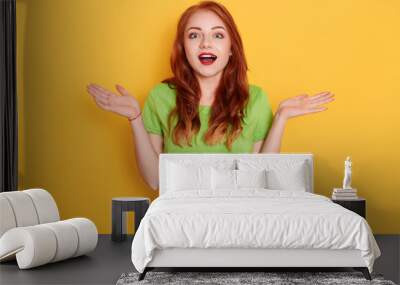 Astonished attractive woman with red hair, spreading palms aside, being shocked to hear news, dressed casual green t shirt, posing isolated over yellow background. Wall mural