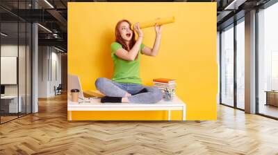 Astonished red haired student female looking through twisting paper aside, has curious facial expression, keeps mouth opened, young girl sitting on desk near laptop and books. Wall mural