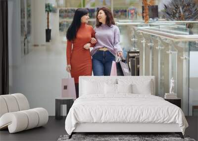 Attractive happy young women in shopping mall walking and smiling, holding shopping bags, spending time together, buying new clothing, expressing positive emotions. Wall mural