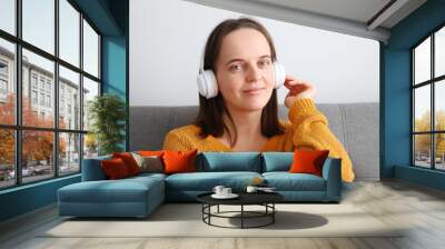 Beautiful attractive cute relaxed woman listening to music with headphones sitting on sofa at home enjoying favorite playlist looking at camera with smile Wall mural