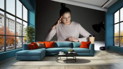 Beautiful student sits at white desk against dark background, tries to concentrate under light of lamp, wears casual jamper and optical glasses, keeps hand on temple, prepares for examinations. Wall mural