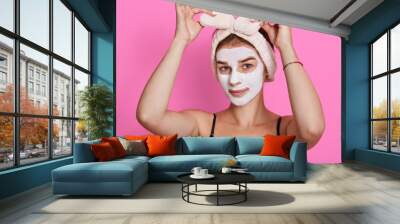 Beautiful woman with white clay facial mask on face, touching her hairband and looking directly at camera, beauty posing isolated over rosy background. Wall mural