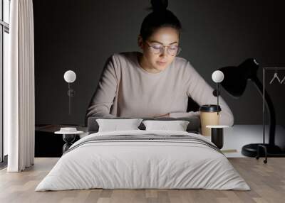 Busy female teenager reads book, uses table lamp, prepares for final examination, sits at desktop, wears optical round glasses, poses against dark background. Reading in evening. Studying concept Wall mural