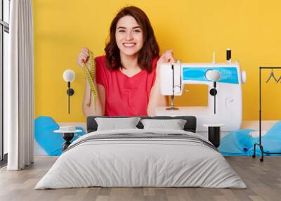 Close up portrait of charming woman seamstress sitting at table with sewing machine on yellow background in studio, dressmaker sews new dress, looks happy, holds measure tape, designer makes outfit. Wall mural