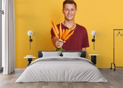 Close up portrait of handsome man holding bunch of fresh carrots isolated over yellow background, attractive guy wearing maroon casual t shirt posing with vegetables in hands, looks happy. Wall mural