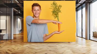 Close up portrait of young attractive caucasian man holding carrot over yellow background, male wearing gray t shirt posing in studio, guy looking smiling directly at camera. Healthy eating concept. Wall mural