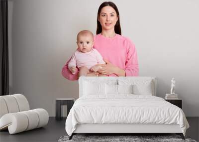 Closeup portrait of beautiful dark haired woman wearing pink sweatshirt and pants holding on hands her little daughter on white background. Family, love, lifestyle, motherhood and tender moments. Wall mural