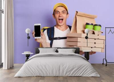 Courier man wearing white T-shirt with thermo backpack holding pizza boxes and coffee in disposable cups isolated over purple background, showing cell phone with empty display for advertisement. Wall mural