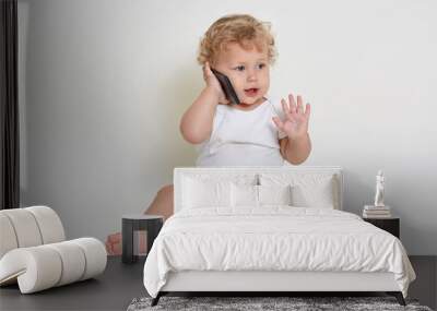 Cute and joyful baby boy playing talking with somebody via smartphone while sitting indoors on floor, looking away and raising palm up, wearing white body suit, play with mobile phone. Wall mural