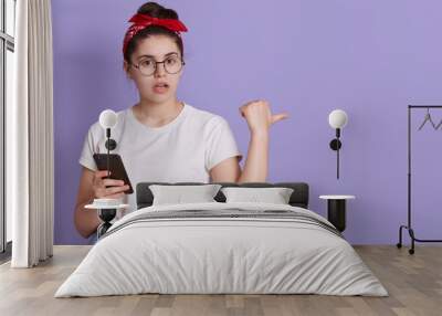 Cute young girl white t-shirt posing against lilac background, holding in hand mobile phone, pointing fingers aside on copy pace, astonished lady with cellphone. Wall mural