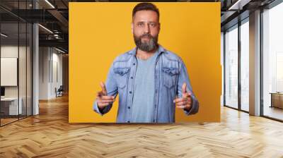 Discreet handsome bearded man weaing light blue t shirt, jeans jacket, showing direction in front of himself with two forefingers. Middle aged confident model posing isolated over yellow background. Wall mural