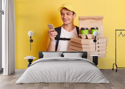 Fast service and best food delivery. Smiling Caucasian delivery man wearing cap and white t shirt holding pizza boxes and takeaway coffee and using mobile phone, isolated on yellow background. Wall mural