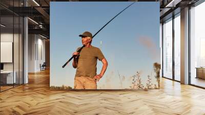 Fisherman posing with fishing rods in hands with blue sky on background, being ready to catch fish, looking pensive side, wearing green t shirt and short. Wall mural