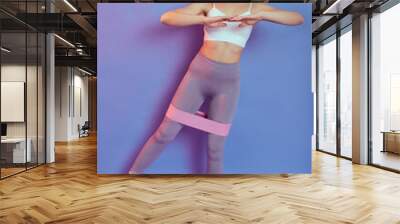 Fitness girl doing exercises while standing indoor, raises leg for hip abductor activation, workout with rubber strap elastic, looks at camera, wears stylish sportswear, keeps hands in front of chest. Wall mural