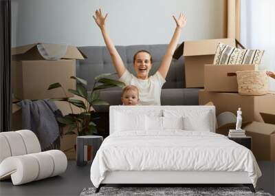 Full length portrait of excited satisfied happy woman wearing white t shirt sitting on floor near sofa with raised hands, rejoicing to relocate to a new house, holding her cute kid on legs. Wall mural