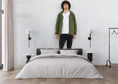 Full length portrait of slim curly haired young man posing over white wall outdoors, looking directly at camera, standing straight, wearing casual clothes, street style. People and style concept. Wall mural