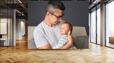 Handsome Caucasian dad holding newborn daughter smiling and caring for baby enjoying precious childhood moments while caring for newborn son sitting on couch Wall mural