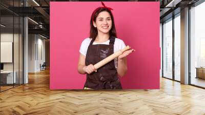 Happy chef woman holding rolling pin in hands isolated over pink background, smiling housewife wearing brown apron, white shirt and red head band looking at camera, female being in good mood. Wall mural