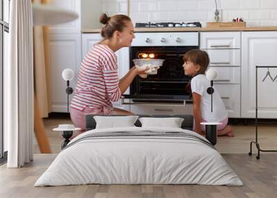 Happy young mum and cute kid daughter baking pie or cake in kitchen, mother and child smelling delicious tasty dessert, people sitting on floor, enjoying flavor. Wall mural