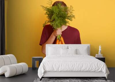 Horizontal shot of attracvtive man holding bunch of fresh carrots, young model isolated over yellow background, handsome guy wearing maroon casual t shirt, covering his face with vegetables. Wall mural