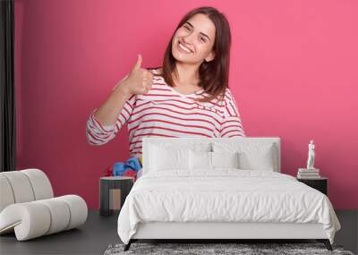 Horizontal shot of happy brunette woman showing thumb up and looking directly art camera, wearing casual attire, holding box with clothes for secondary using, female volunteer helping poor people. Wall mural