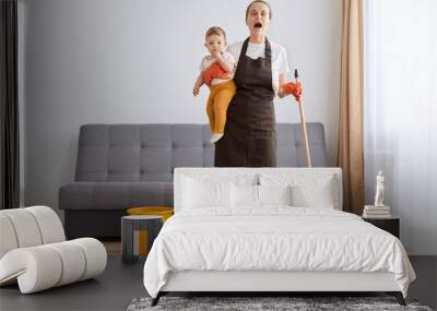 Horizontal shot of little baby girl in young mother hands, desperate woman cleaning at home, tired exhausted mom mopping floor, tidying together in living room. Wall mural