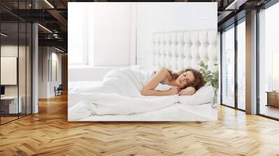 Horizontal shot of relaxed carefree female under white bedclothes in bed at bedroom, sees pleasant dreams, keeps eyes closed, enjoys good rest at home and calm atmosphere. People and rest concept Wall mural