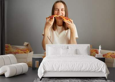 Hungry funny woman with brown hair wearing white T-shirt sitting at table among fast food isolated over gray background holding two slices of pizza eating junk food. Wall mural