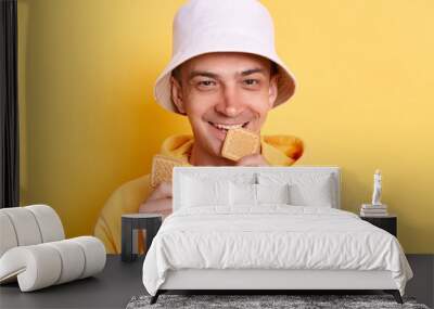 Hungry positive Caucasian young adult man wearing casual style hoodie and panama posing isolated over yellow background, holding and biting cookies, looking at camera with happiness. Wall mural