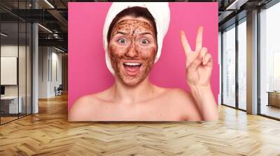 Image of adorable magnetic half naked lady raising two fingers, opening her mouth in sincere smile, posing with brown mask on her face, keeping her face in good condition. Beauty and care concept. Wall mural