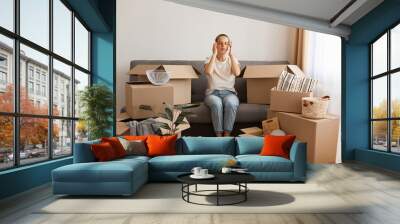 Image of attractive tired woman wearing white t shirt and jeans sitting on sofa during relocating to a new apartment, being exhausted and trying to concentrate. Wall mural