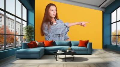 Image of european woamn wearing casual clothes looking far away with opened mouth and points with her fore finger, looks astonished, posing isolated over yellow background. People lifestyle concept. Wall mural