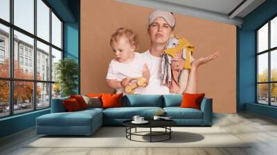 Image of young mother with little child in arms, spreading arm aside, having tired upset facial expression, female being exhausted, has lot of work, posing isolated over brown background. Wall mural