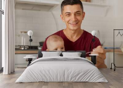 Indoor portrait of happy young father wearing maroon t shirt holding baby on his knees and feeding son or daughter with vegetable or fruit puree in kitchen. Wall mural