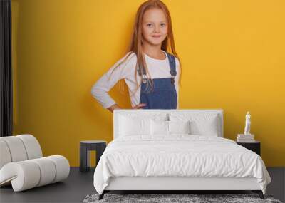 Indoor shot of cute little girl wearing jeans overall and white shirt, posing in studio isolated over yellow background, keeps hand on hip, looks directly at camera, charming girl has blonde hair. Wall mural