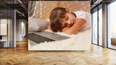 Indoor shot of sleepy little dark haired girl wearing white t shirt or pajama laying on floor in wigwam with closed eyes, falling asleep while watching cartoons in peetee tent. Wall mural