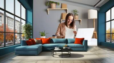 Indoor shot of smiling young adult beautiful brown haired woman wearing beige jacket posing in office, working on laptop and and with paper documents, enjoying her work. Wall mural