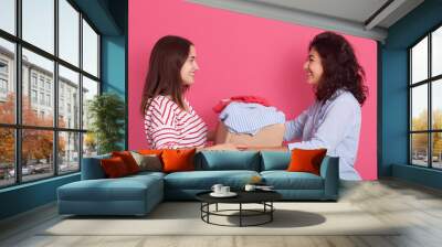 Indoor shot of two attractive women posing isolated over rosy background, smiling lady handing big carton box with clothes, young female gives charity clothing for poor people. Donation concept. Wall mural