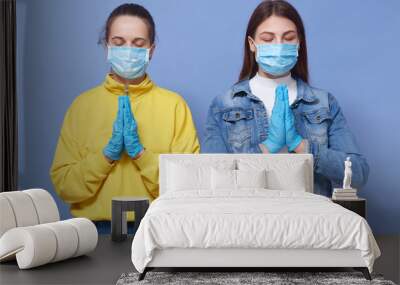 Indoor shot of two calm attractive females standing isolated over blue background in studio, keeping hands together, closing eyes, wearing masks, praying, fighting with coronavirus. Health concept. Wall mural