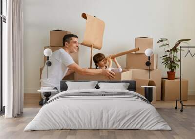 Indoor shot of young brunette man wearing casual attire sitting on floor and playing with his cute dark haired daughter, little girl sitting on a ship made from carton box and looking in spyglass. Wall mural