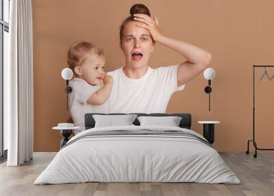 Indoor shot of young tired shocked mother holding a child in her arms, female wearing white casual style T-shirt posing with a baby isolated on brown background, screaming, keeps hand on head. Wall mural