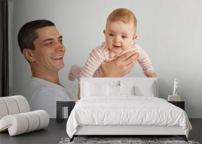 Laughing brunette man wearing white casual t shirt holding his infant daughter, posing at home in light room, father throwing kid in the air, pretending child is flying. Wall mural