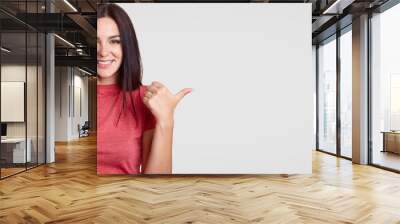 Optimistic young female with toothy friendly smile points aside with thumb, shows blank space for your advertising content or promotional text, has long dark hair, isolated over white background Wall mural